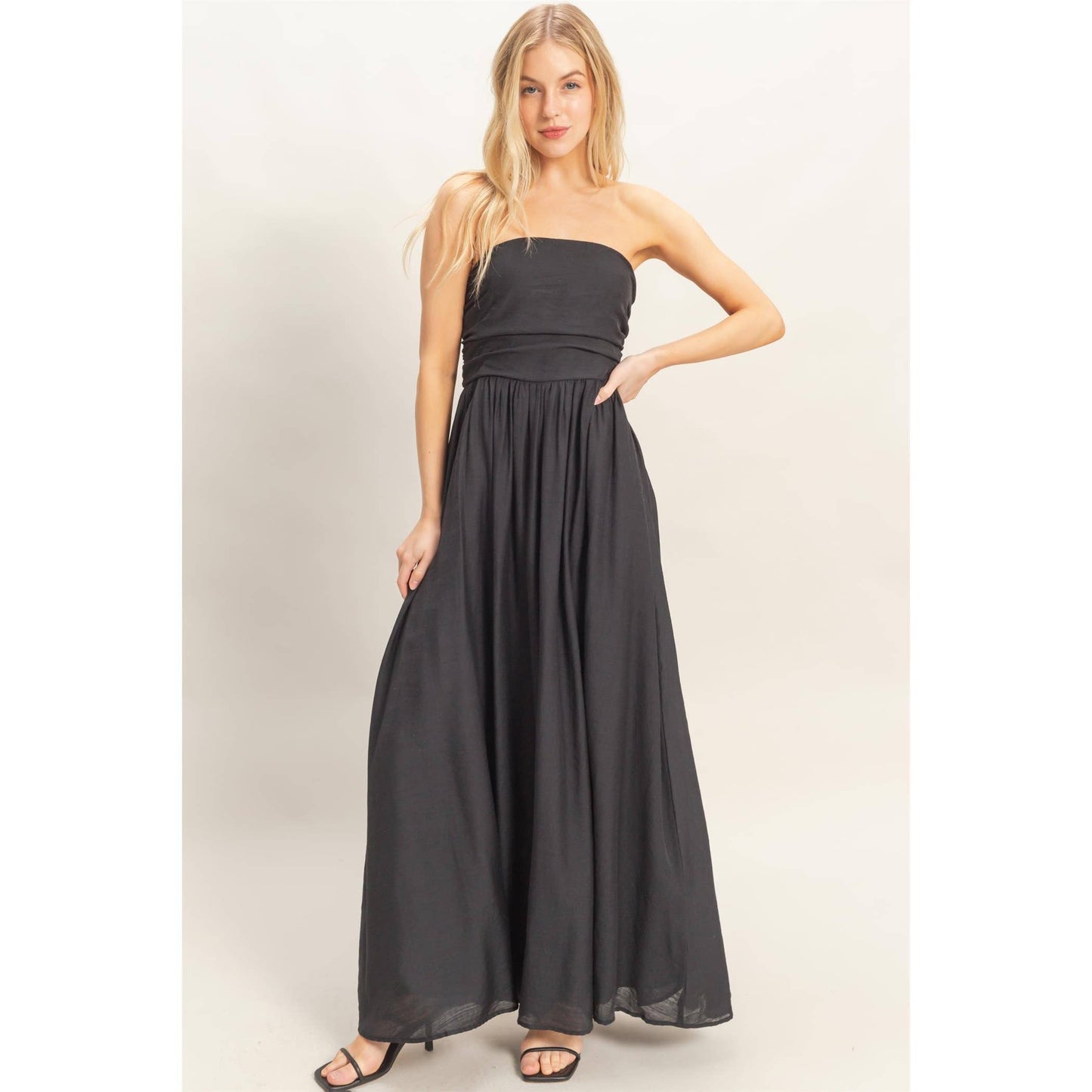 Strapless black smocked dress with a tie-back sash, flowing maxi length on a model with sandals.