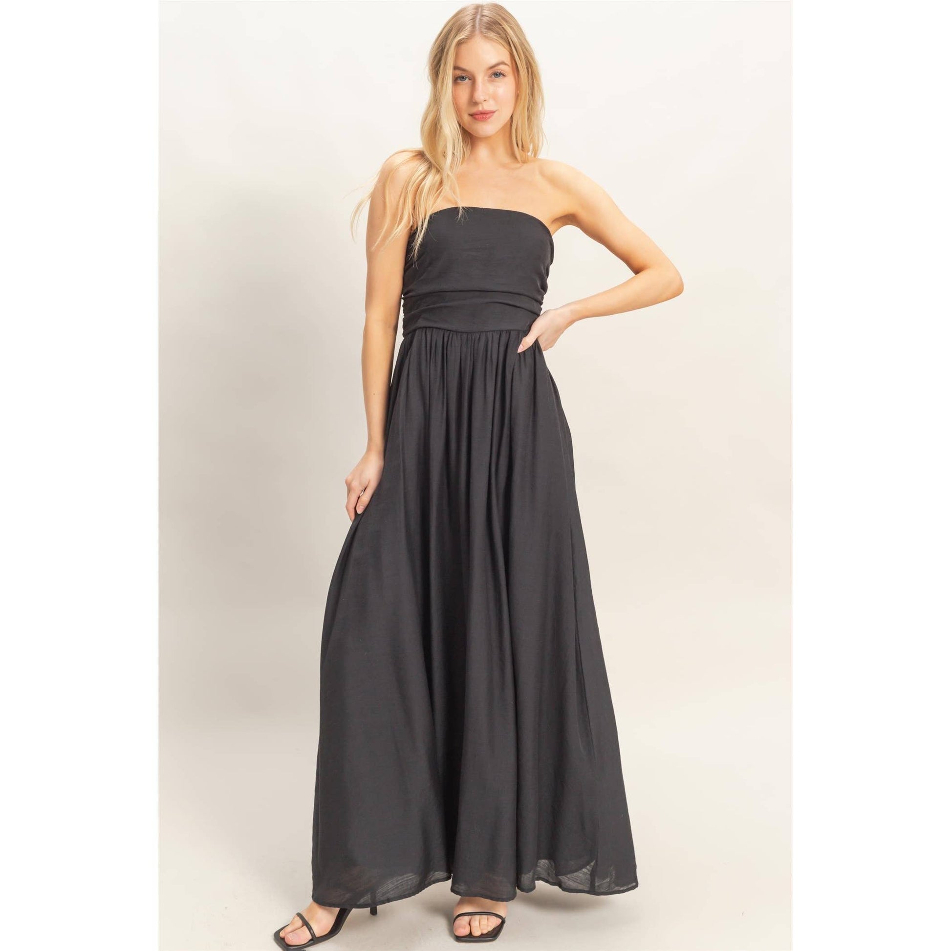 Strapless black smocked dress with a tie-back sash, flowing maxi length on a model with sandals.